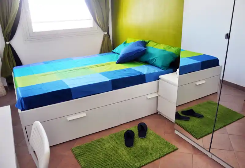 Dormitorio luminoso y ordenado con cama bien visible, ropa de cama colorida y alfombras verdes a juego. La luz natural entra por la ventana y un armario con espejo amplía visualmente la habitación.