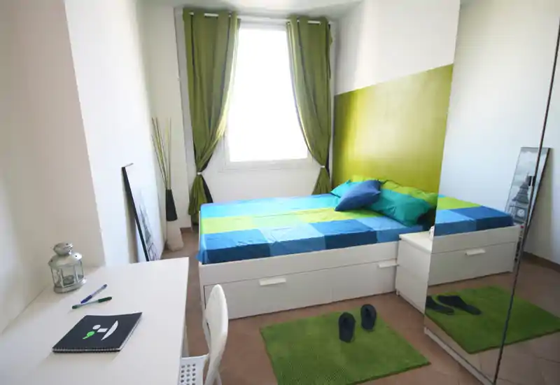Dormitorio moderno y luminoso con cama doble, ropa de cama azul y verde, cojines a juego y cortinas verdes. Cama con almacenaje blanco y armario con espejo que optimizan el espacio, con luz natural desde la ventana.