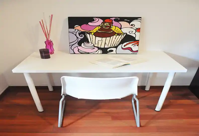 Piccolo angolo per lavorare/sedersi con scrivania bianca, sedia, vaso decorativo e quadro colorato — stile pulito e semplice.