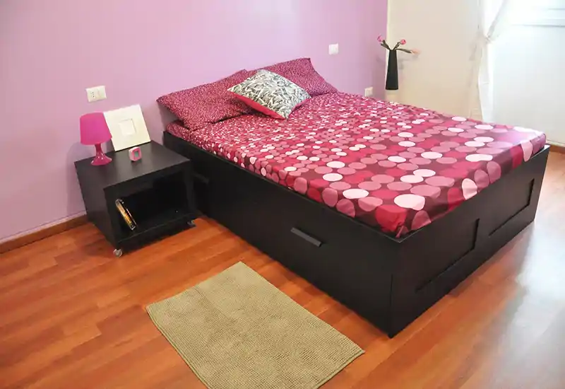 Camera da letto luminosa e ben arredata con letto matrimoniale con biancheria rosa a fantasia, comodino con lampada e finestra che illumina la stanza.