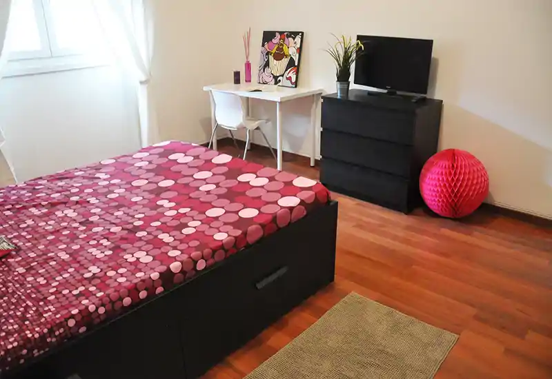 Camera da letto luminosa con un ampio letto matrimoniale con copriletto a fantasia, una piccola scrivania vicino alla finestra e un comò con TV — moderna, accogliente e ben illuminata.