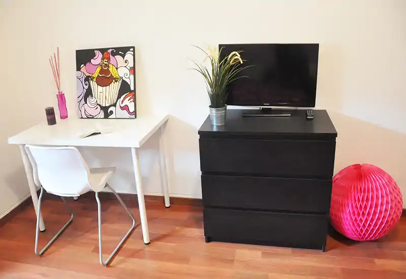 Angolo living arredato in modo compatto con scrivania bianca e sedia, cassettiera nera a tre cassetti con TV e pianta decorativa; pavimento in legno e accenti colorati creano un ambiente ordinato e accogliente.
