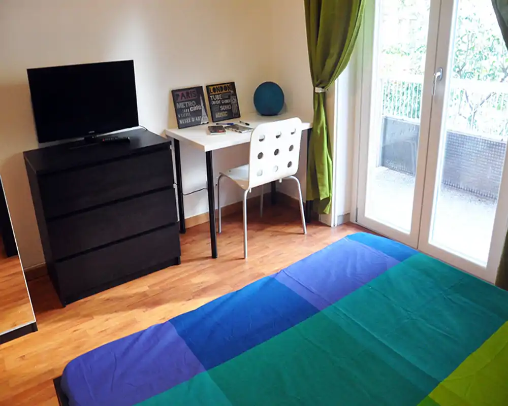 Dormitorio ordenado y luminoso con colcha colorida, pequeño escritorio y silla junto a la puerta del balcón — ideal para una persona o estudiante.