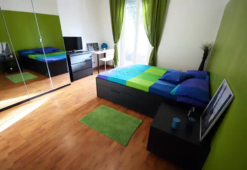 Dormitorio luminoso y bien decorado con cama doble, pared verde y cortinas a juego. Armario con espejo, suelo de madera y un pequeño escritorio junto a la ventana crean un ambiente fresco y ligero.