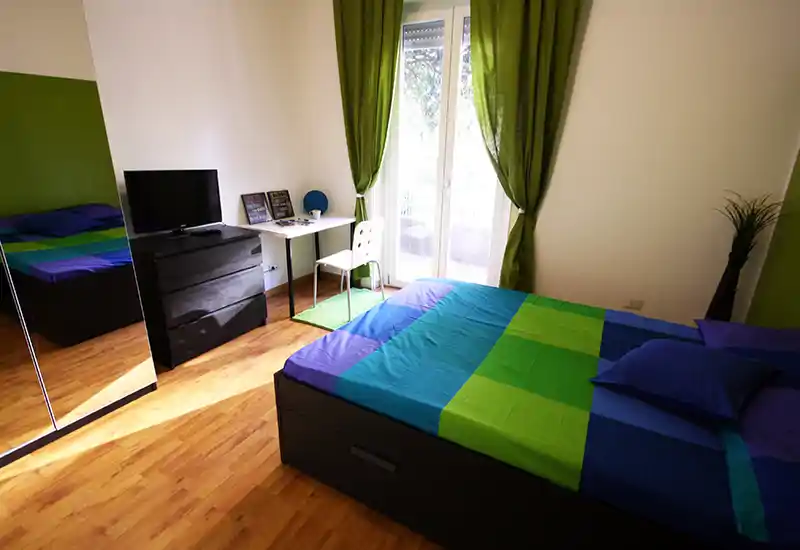 Dormitorio moderno y luminoso con cama doble vestida con ropa de cama azul y verde, suelo de madera, armario con espejo, televisor sobre cómoda y un pequeño escritorio junto a la puerta del balcón que aporta luz natural.