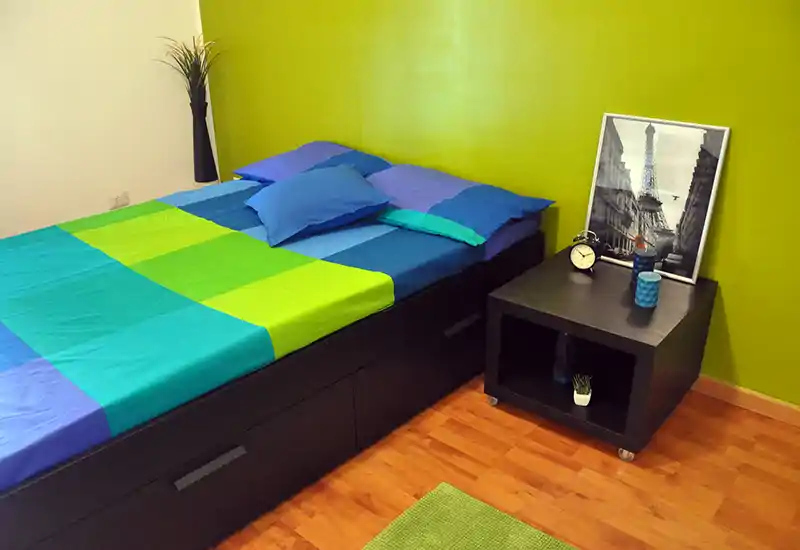Dormitorio luminoso y bien arreglado con una cama grande vestida con ropa de cama azul y verde brillante, mesita cúbica con decoración y un cuadro enmarcado, suelo de madera — ideal para anuncios de alquiler.
