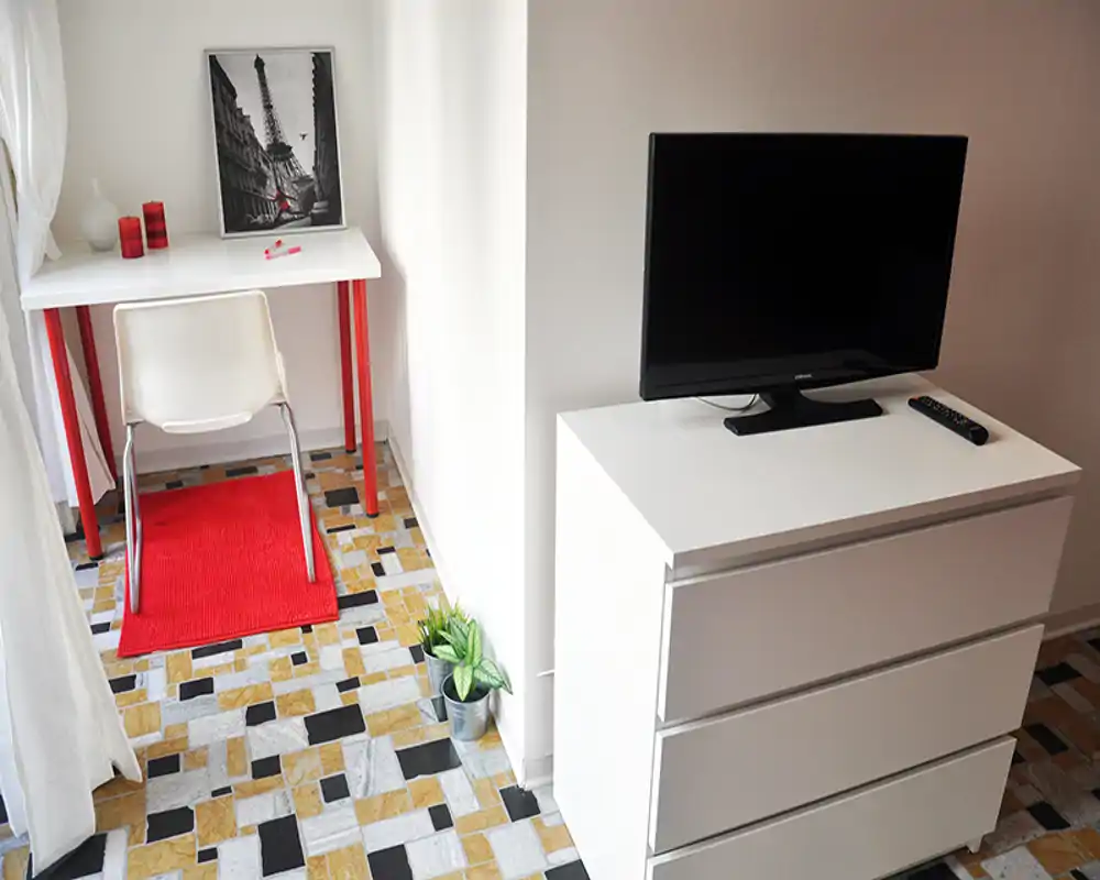 Area giorno compatta con cassettiera bianca e TV, una piccola scrivania con sedia e accenti rossi decorativi; il pavimento a motivi e la luce naturale rendono lo spazio gradevole e funzionale.