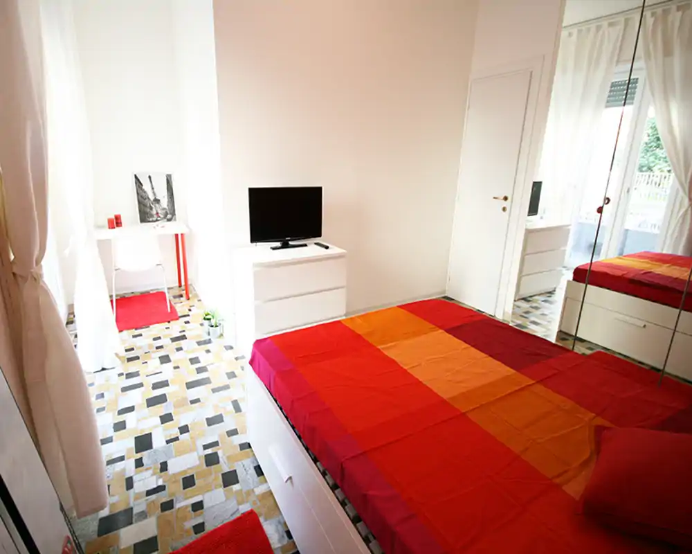 Camera da letto luminosa e colorata con un ampio letto matrimoniale con copriletto rosso-arancione, armadio con specchi, comò con TV e una piccola scrivania vicino alla finestra — ideale per un affitto soleggiato e accogliente.