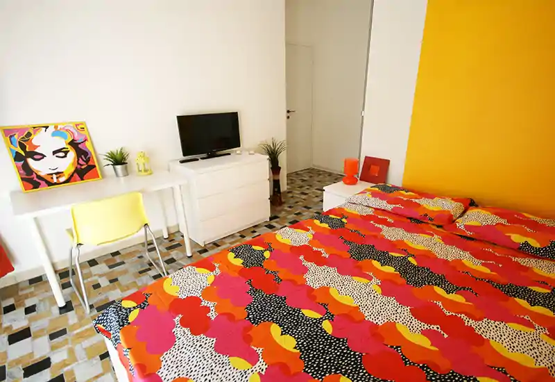 Camera da letto luminosa e colorata con un grande letto decorato in primo piano, una scrivania bianca con sedia gialla e una piccola TV su una cassettiera; stile contemporaneo e luce naturale abbondante.