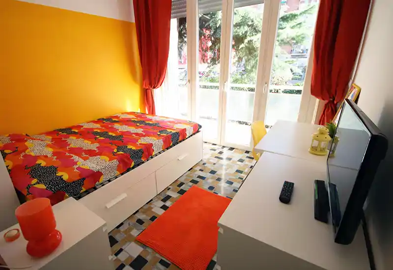 Camera singola luminosa con biancheria colorata, letto contenitore bianco, scrivania e ampie porte-finestre che lasciano entrare molta luce naturale — perfetta per un affitto vivace.