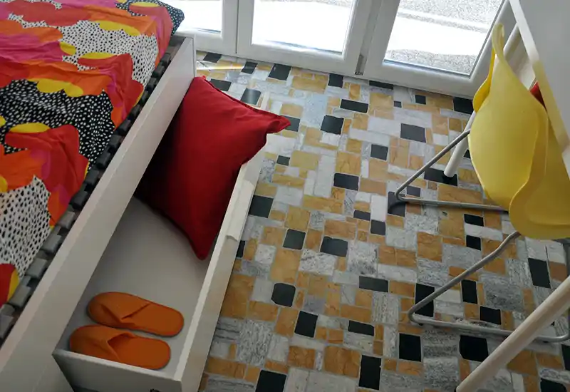 Angolo camera con un letto singolo dotato di copriletto colorato e cassettone estraibile; luce naturale dalle porte-finestre e pavimento piastrellato creano un'atmosfera vivace.