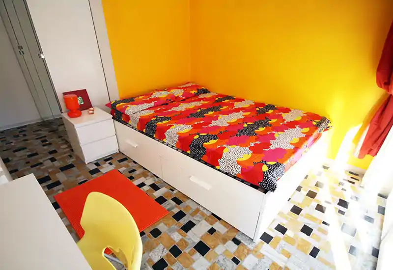 Camera singola luminosa e colorata con letto contenitore bianco, copriletto fantasia vivace e pareti gialle calde. La luce naturale rende l’ambiente accogliente e perfetto per un singolo.