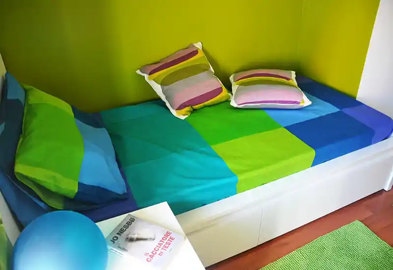 Camera singola compatta con letto colorato ben rifatto, cuscini decorativi e arredi luminosi e freschi — ideale per affitti per studenti o ospiti.