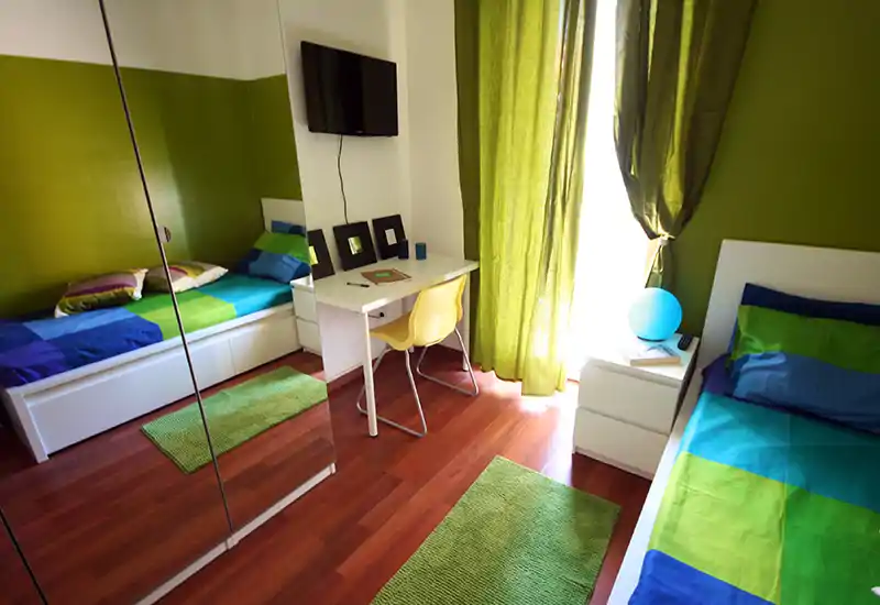 Camera da letto doppia luminosa e moderna con biancheria colorata, tende verdi e tappeti abbinati; dispone di scrivania, comodino e armadio con ante a specchio che aumentano la profondità.