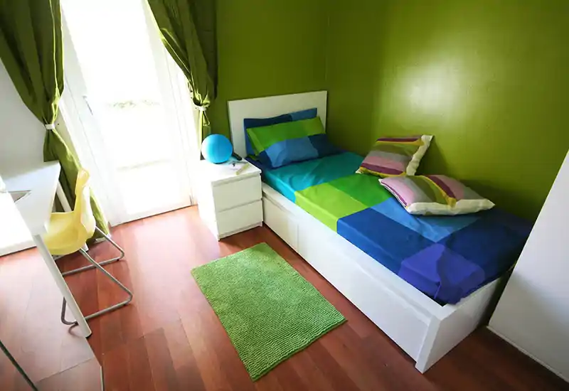 Camera singola compatta e luminosa con letto colorato, cuscini coordinati e un piccolo comodino. La luce naturale dalla porta finestra e i toni verdi creano un ambiente fresco e accogliente.