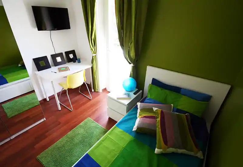 Camera da letto moderna e luminosa con letto singolo con biancheria colorata, comodino con lampada, piccolo scrittoio e TV a parete; pareti verdi e luce naturale dalla porta donano un'atmosfera allegra.