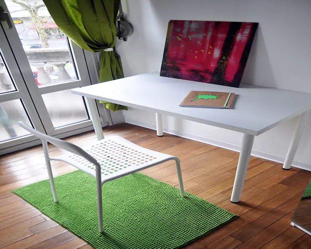 Area di lavoro minimalista e luminosa con scrivania bianca e sedia traforata accanto a una grande finestra; tappeto e tenda verdi aggiungono colore e un piccolo quadro appoggiato al muro.