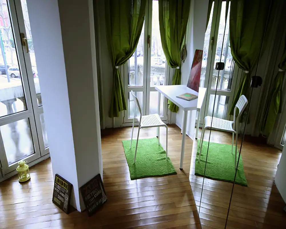 Angolo luminoso con ampie finestre, un piccolo tavolo bianco e due sedie su tappeti verdi; zona living minimal e moderna ideale per colazione o lavoro.