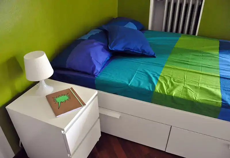 Angolo camera luminoso e ordinato con letto completo con biancheria blu e verde, comodino bianco con lampada e quaderno, e termosifone contro una parete verde lime.