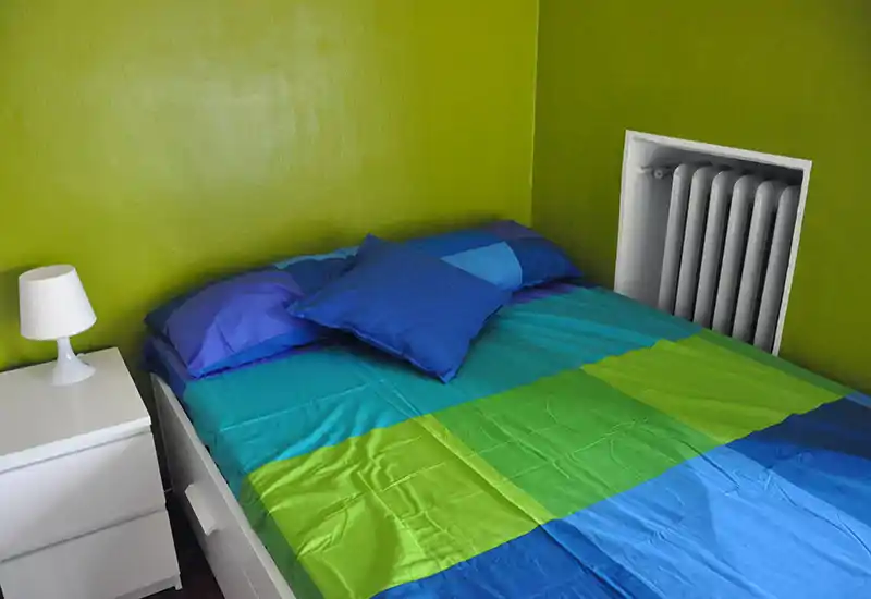 Camera da letto luminosa con biancheria colorata blu-verde, cuscini e comodino bianco con lampada; ambiente moderno, semplice e ben illuminato, perfetto per un annuncio.