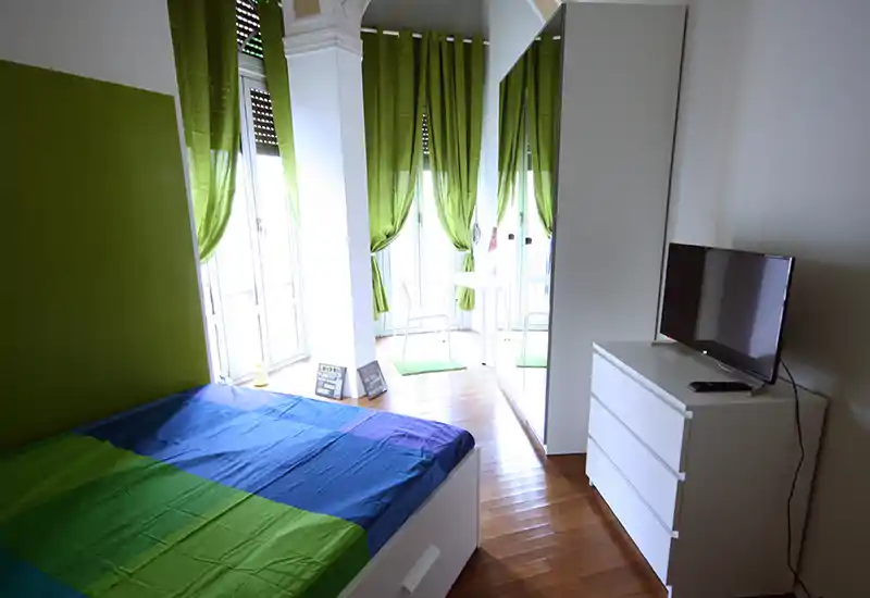 Camera da letto luminosa e moderna con un ampio letto in primo piano, tende verdi e una cassettiera bianca con TV; pavimento in legno lucido e armadio con specchio ampliano lo spazio.