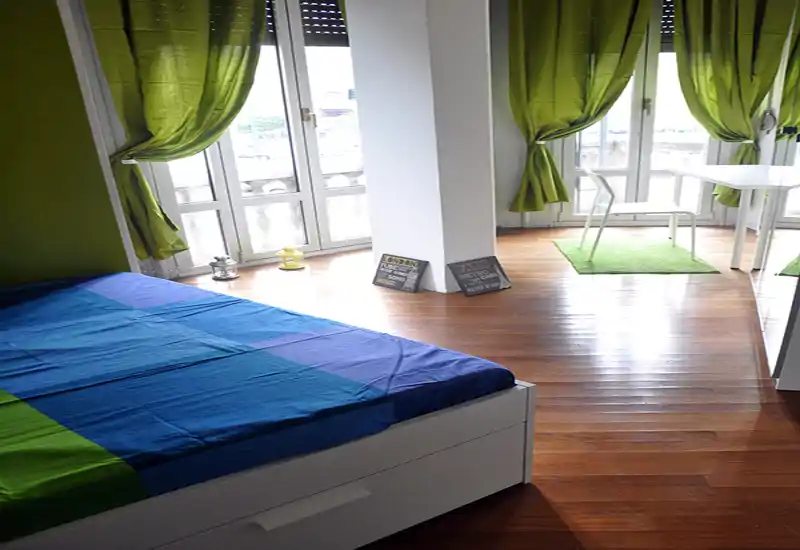Camera da letto luminosa e moderna con letto visibile, ampie finestre con tende verdi e pavimento in legno che creano un'atmosfera fresca e ariosa.