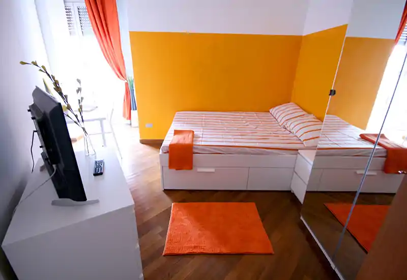 Bright, modern bedroom with a white platform bed, orange accents and natural light from a window; mirrored wardrobe adds depth.