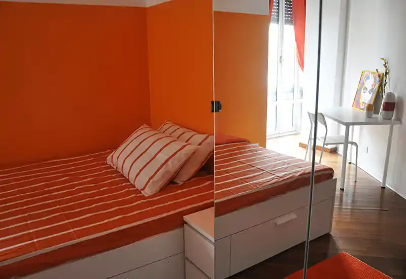 Bright, modern bedroom with an orange color scheme, striped bedding and a mirrored wardrobe that adds depth. A small desk sits near the window, making the space practical for work or study.
