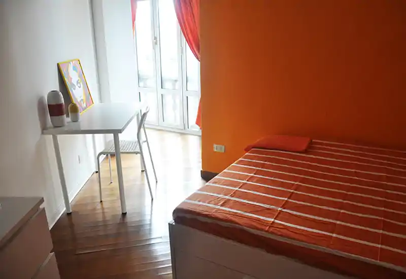 Bright, modern bedroom with an orange accent wall and a neatly made bed; natural light fills the room from a large window and a small desk provides a functional workspace.