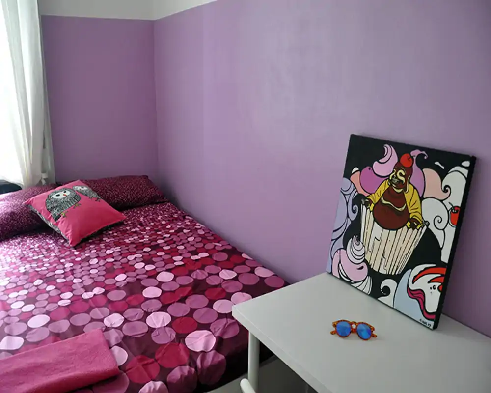 Dormitorio luminoso y acogedor con una cama doble estampada junto a una pared lila, almohada decorativa y una mesita con una obra de arte — perfecto para un anuncio de alquiler colorido y con estilo.