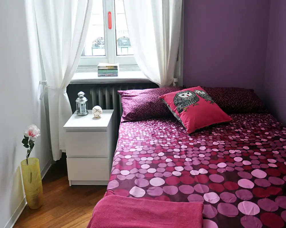Dormitorio luminoso y acogedor con colcha magenta de puntos, cojines decorativos y una mesita de noche blanca bajo una ventana con cortinas.