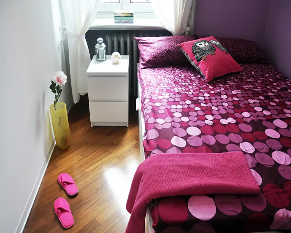 Dormitorio luminoso y acogedor con colcha magenta con estampado, cojín decorativo y mesita de noche bajo una ventana que deja entrar luz natural.