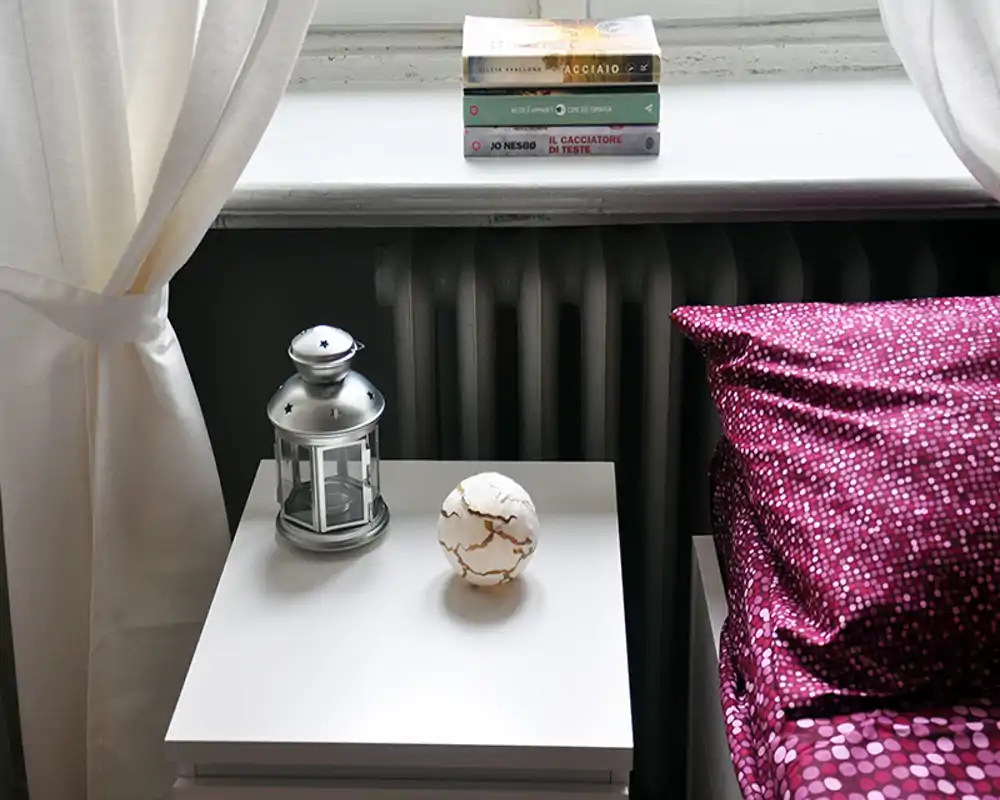 Rincón de dormitorio luminoso y acogedor con mesita, linterna decorativa y cojín magenta estampado junto a la ventana con libros en el alféizar.