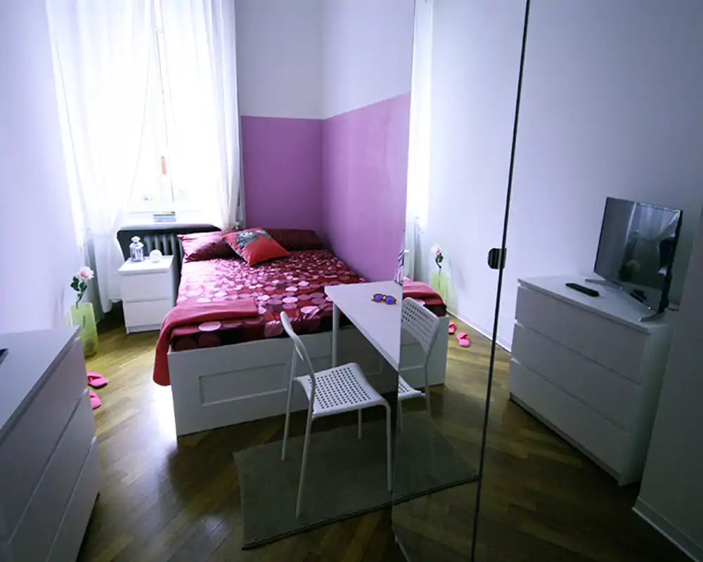 Dormitorio luminoso y compacto con cama doble con ropa de cama roja estampada, mesitas, pequeño escritorio y TV sobre cómoda; mucha luz natural desde la ventana y armario con espejo que aportan sensación de amplitud.