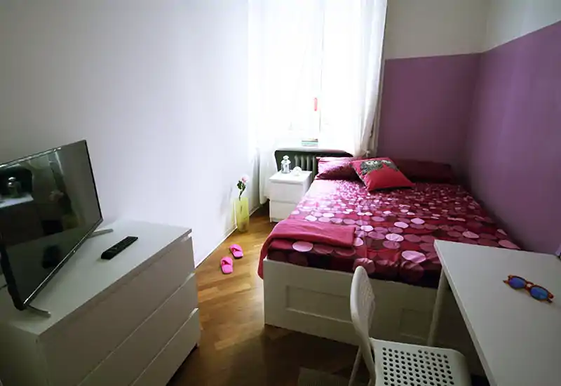 Dormitorio luminoso y ordenado con cama doble con ropa de cama rosa estampada, mesita junto a la ventana, cómoda con TV y un pequeño escritorio — acogedor y listo para huéspedes.