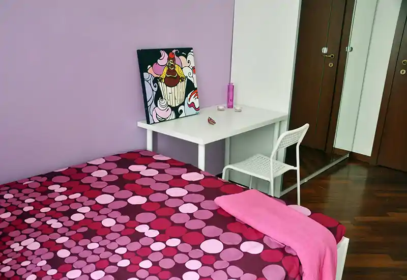 Dormitorio luminoso y con estilo, con colcha rosa y morada de motivos circulares, escritorio y silla blancos y armario con espejo — acogedor y bien coordinado.