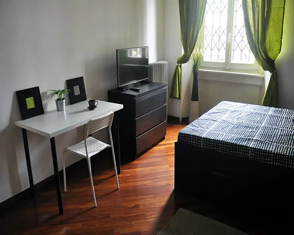 Bright, tidy bedroom with a double bed, wooden floor and a small desk by the wall. Natural light filters through green curtains, and a TV on a chest of drawers adds convenience for guests.