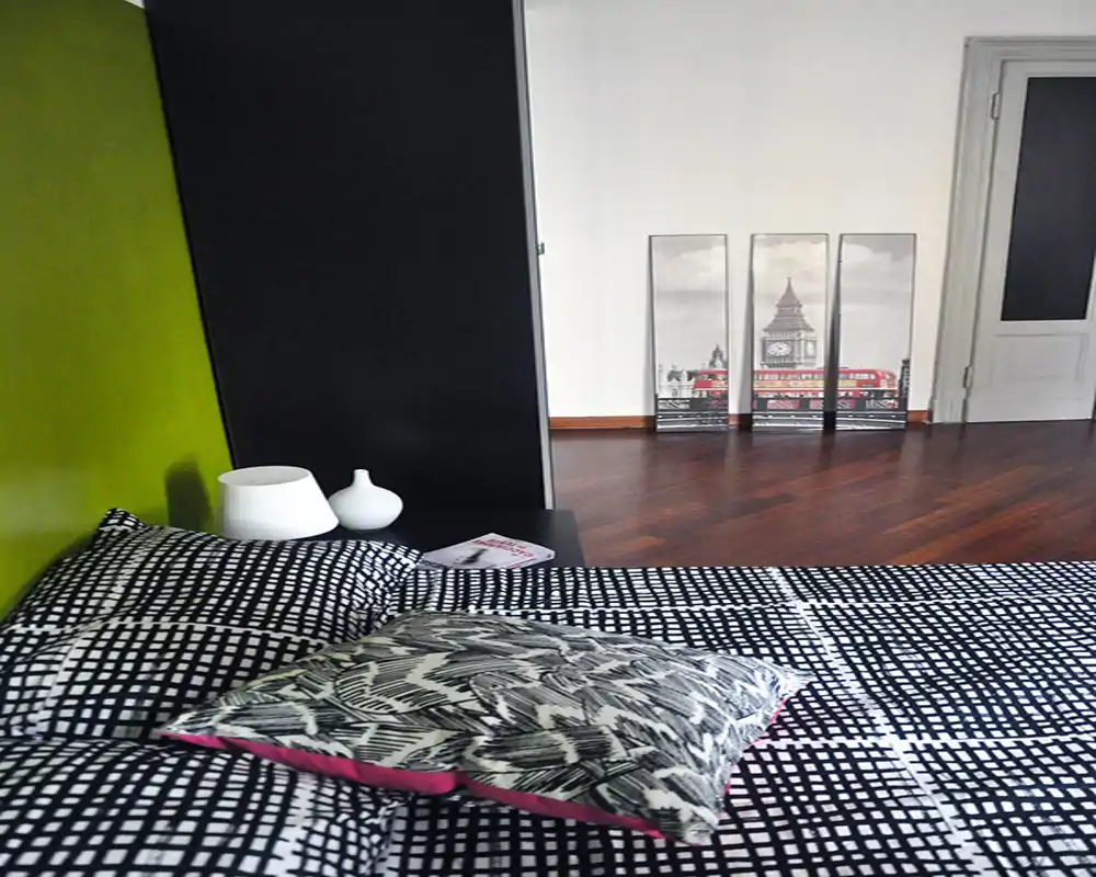 Modern bedroom with a patterned bedspread, decorative cushions and a bedside table with lamp. The room features wooden floors and simple wall art, creating a clean and contemporary atmosphere ideal for rental photos.