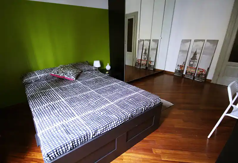 Bright, modern bedroom with a large double bed, patterned bedding, bedside lamp and mirrored wardrobe reflecting the room; warm wooden floors and green accent wall create a contemporary, cozy atmosphere.