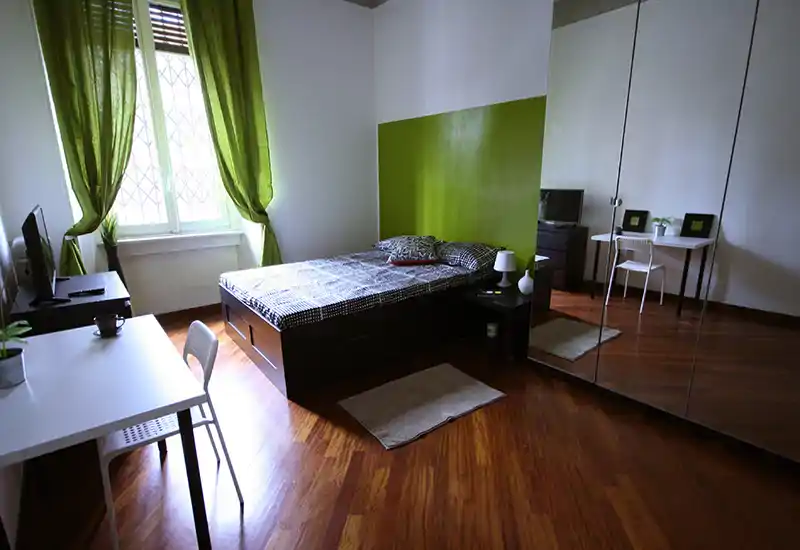 Bright, modern bedroom with a double bed, green accent wall and large mirrored wardrobe. Natural light from a window highlights wooden floors and simple, functional furnishings.
