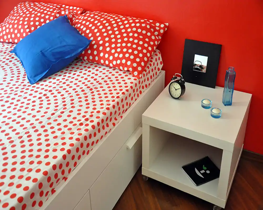 Angolo di camera luminoso e moderno con copriletto a pois, cuscini e un comodino bianco con oggetti decorativi. L’immagine mette in risalto il letto e lo stile del comodino con una parete rossa vivace.