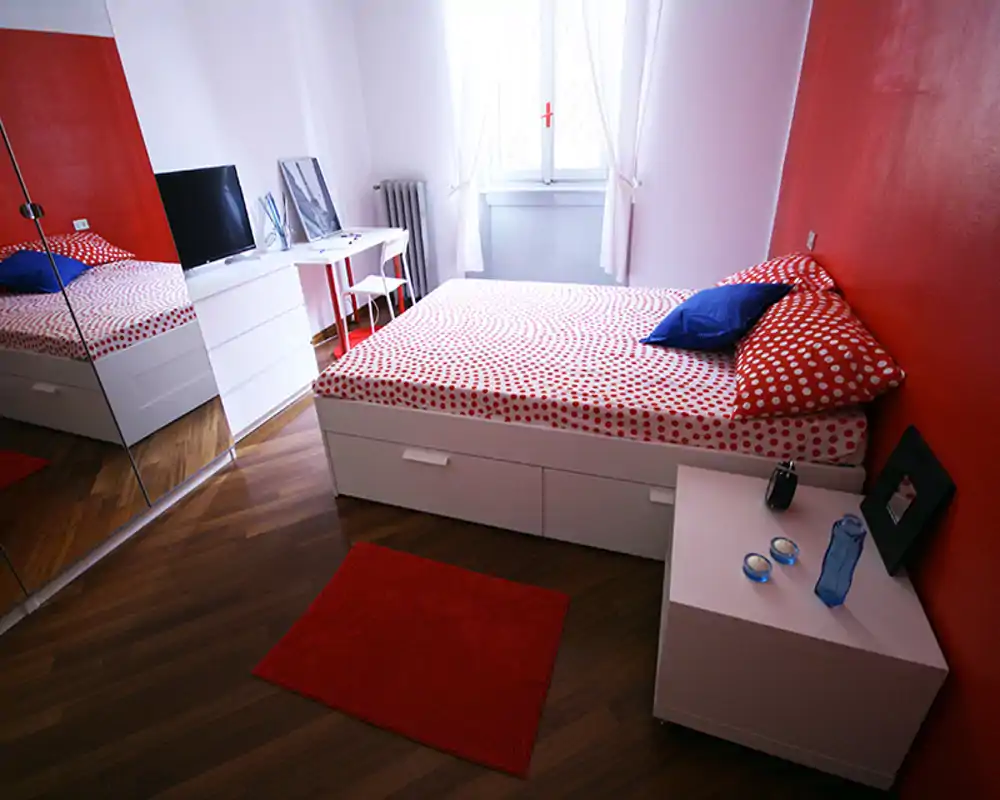 Camera da letto luminosa e moderna con letto matrimoniale con copriletto a pois rossi, cassetti contenitore sotto il letto e grande armadio specchiato che aggiunge profondità — perfetta per un annuncio.