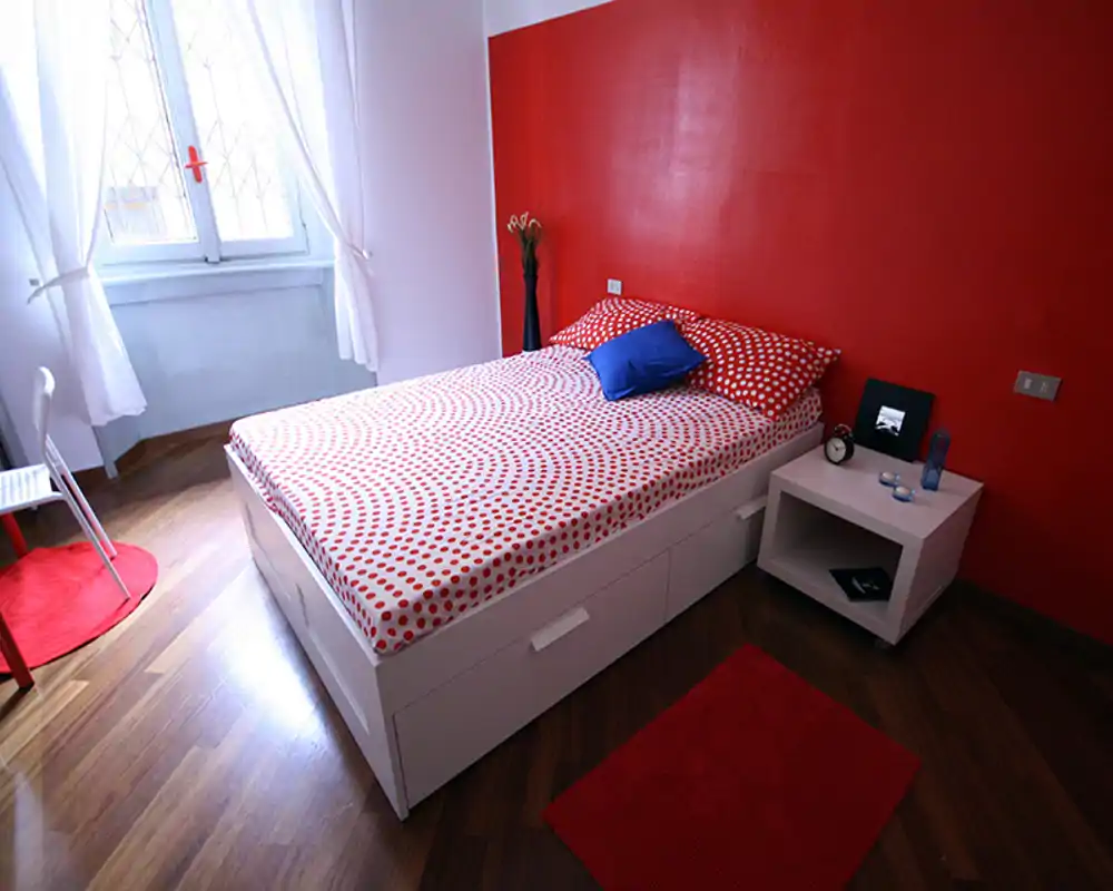 Camera da letto singola luminosa e moderna con letto contenitore bianco, parete rossa a contrasto e biancheria a pois coordinata. Luce naturale dalla finestra e comodino minimal completano uno spazio elegante e ideale per annunci.