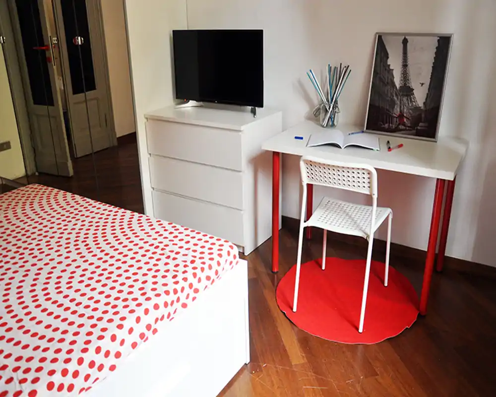 Camera da letto luminosa con copriletto a pois rosso e bianco, comò bianco con TV e una scrivania ordinata con sedia — arredo moderno, semplice e accogliente.