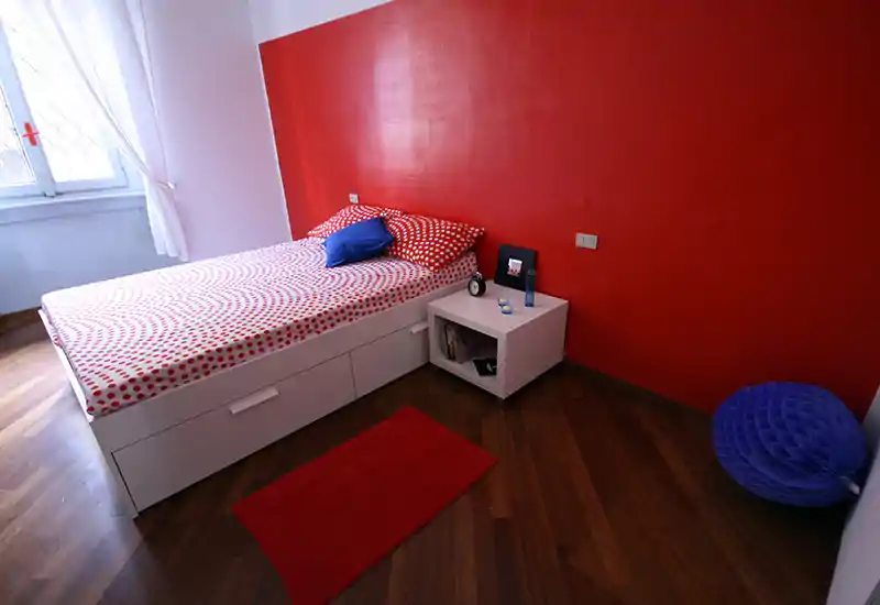 Camera da letto moderna e luminosa con letto bianco con cassetti sotto, biancheria a pois rossi e parete d'accento rossa; luce naturale dalla finestra e un piccolo comodino rendono l'ambiente accogliente.