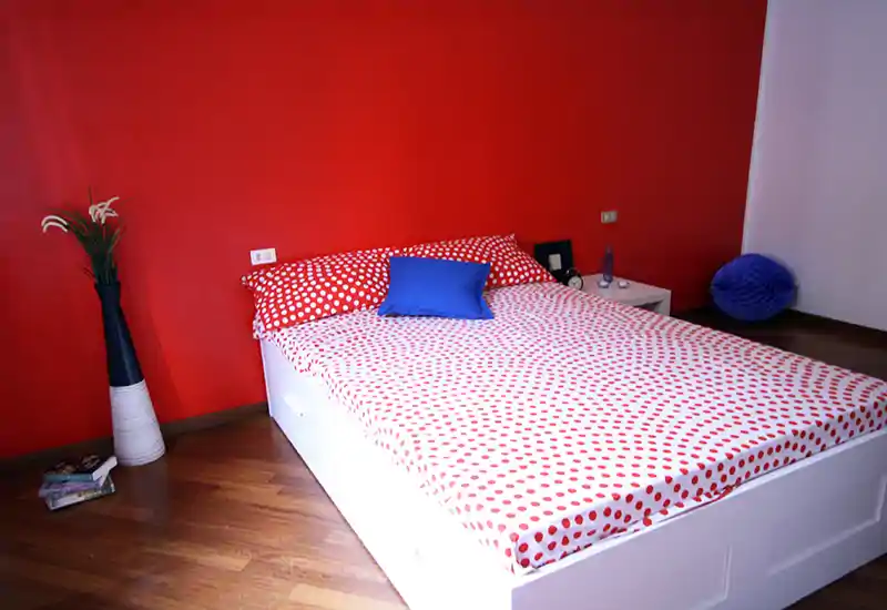 Camera da letto luminosa e moderna con letto bianco su piattaforma, biancheria a pois rossi e cuscino blu a contrasto su una parete rossa intensa.