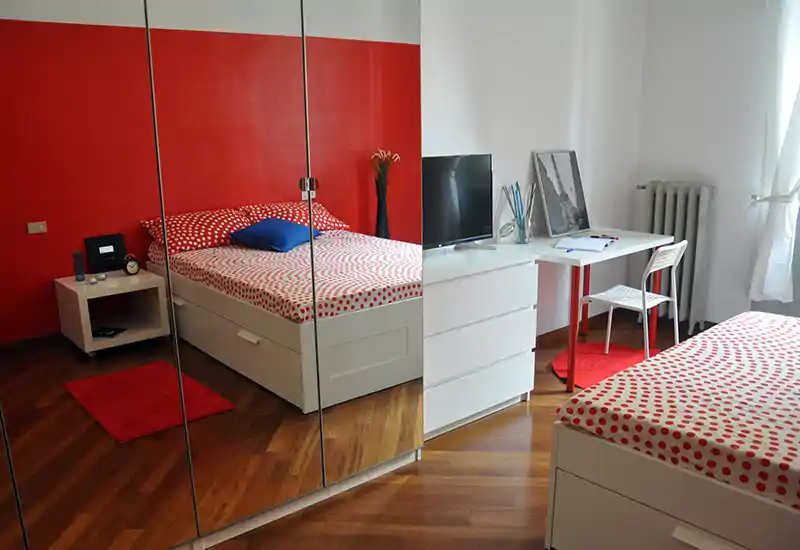 Camera da letto moderna e luminosa con parete rossa e biancheria a pois. Armadio con ante a specchio, cassettiere bianche e una scrivania vicino alla finestra, perfetta per affitti brevi.