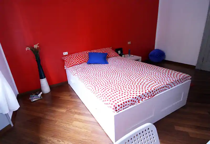 Camera da letto luminosa e moderna con letto contenitore bianco e copriletto a pois rossi su una parete d'accento rossa. Pavimento in legno e arredo minimal creano un'atmosfera fresca e di stile, perfetta per annunci.
