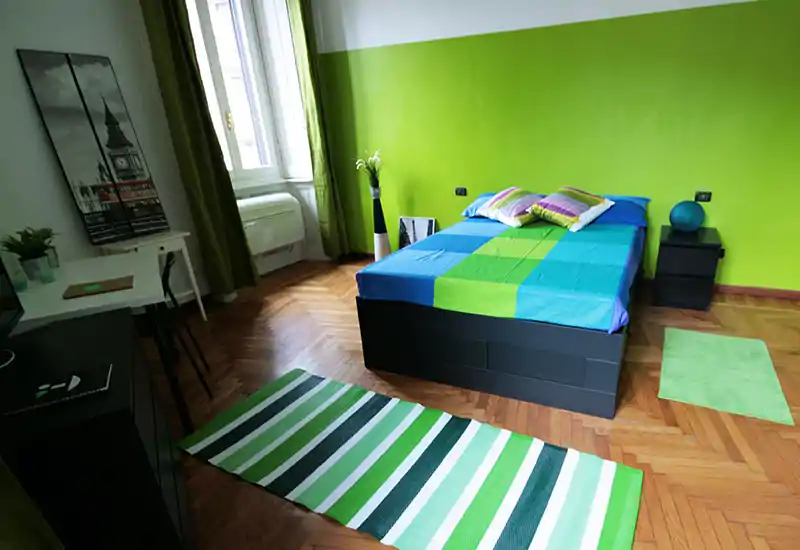 Dormitorio moderno y luminoso con cama doble bien hecha frente a una pared verde intensa, suelo de madera y alfombras a rayas verdes coordinadas.
