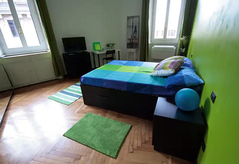 Bright, colorful bedroom with a double bed dressed in blue-green linens, matching rugs and natural light from two windows — well-styled and inviting for short-term rentals.